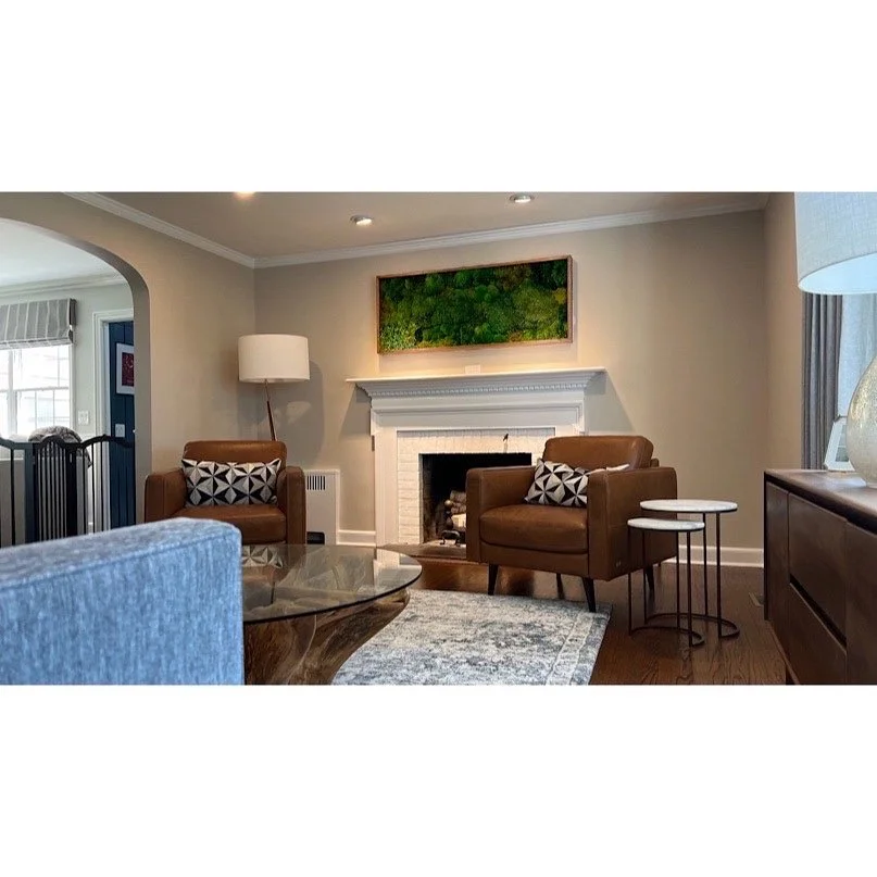 Living room with two brown armchairs with black and white pillows, a white brick fireplace, a framed green landscape above the fireplace, a glass coffee table, a blue armchair, a gray rug, a standing white lamp, a side table with black metal legs, a 