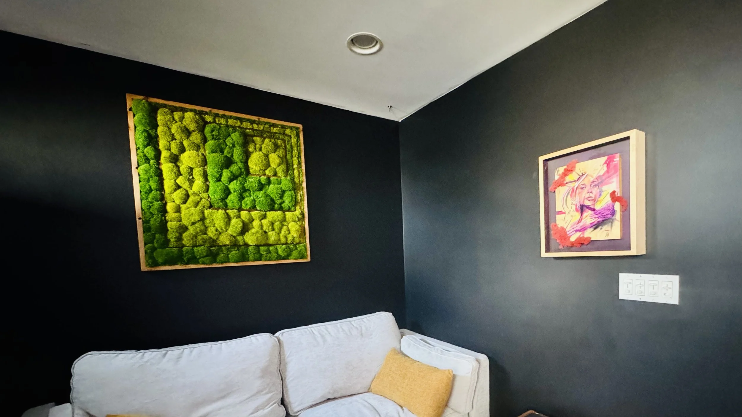 Interior of a living room with a dark wall, white sofa with a yellow pillow, framed green moss art on the left wall, and a framed colorful portrait on the right wall.
