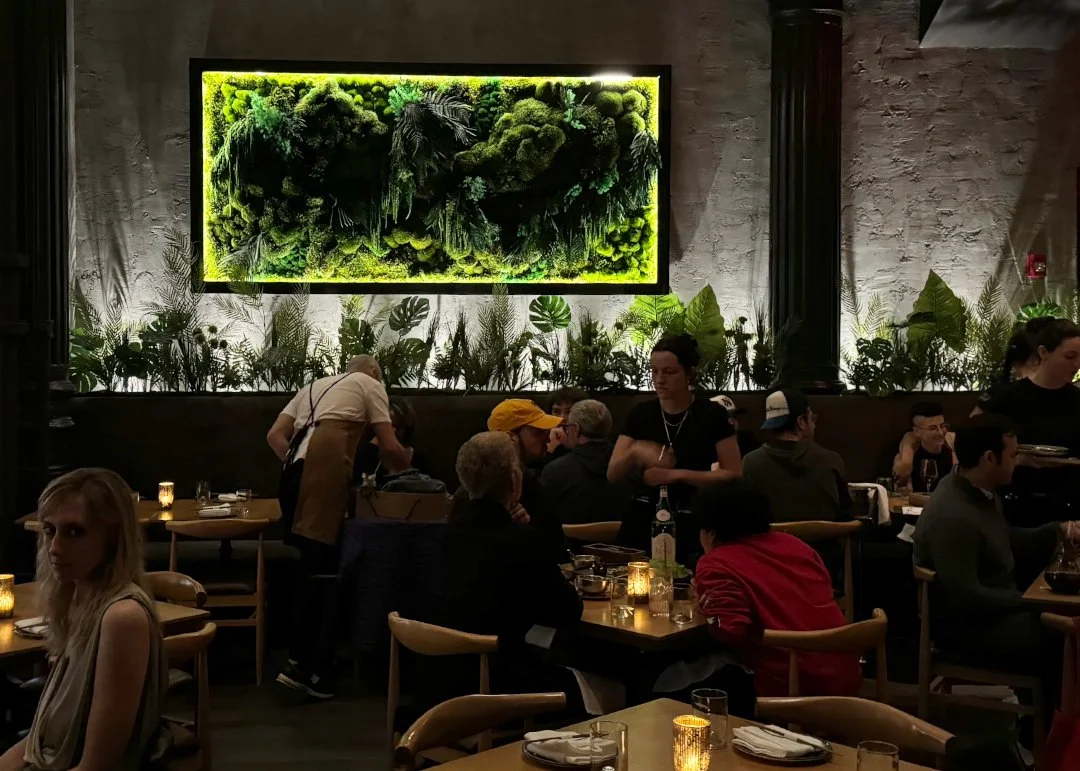 Interior of a restaurant with a green plant art installation on the wall and people dining at tables.