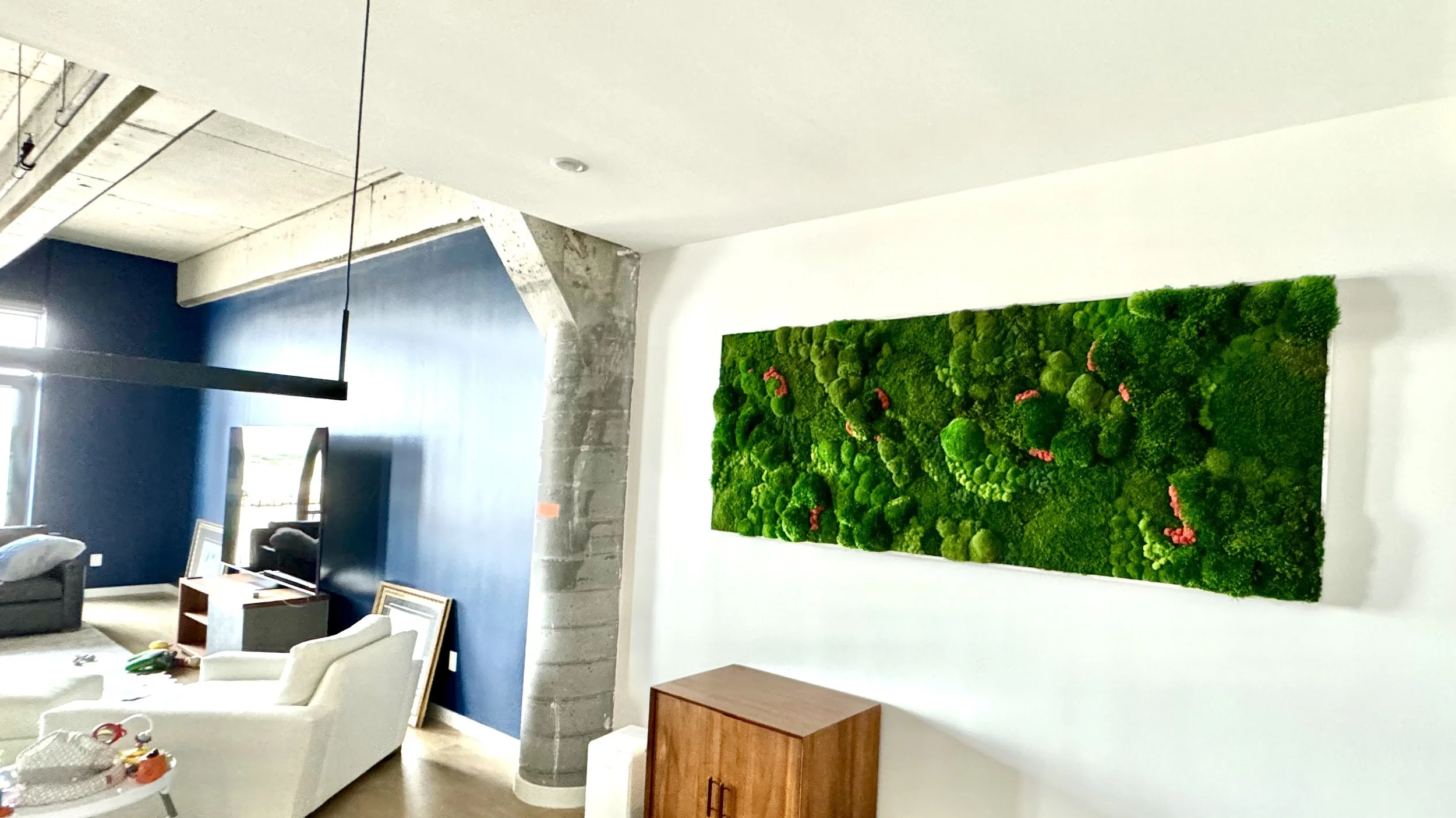 Living room with blue accent wall, green moss wall art, white seating, wooden cabinet, and a flat-screen TV.
