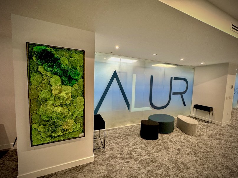 Lobby or waiting area with a glass wall featuring the word AUR, seating options including a black chair and circular ottomans, and a green moss wall decor.