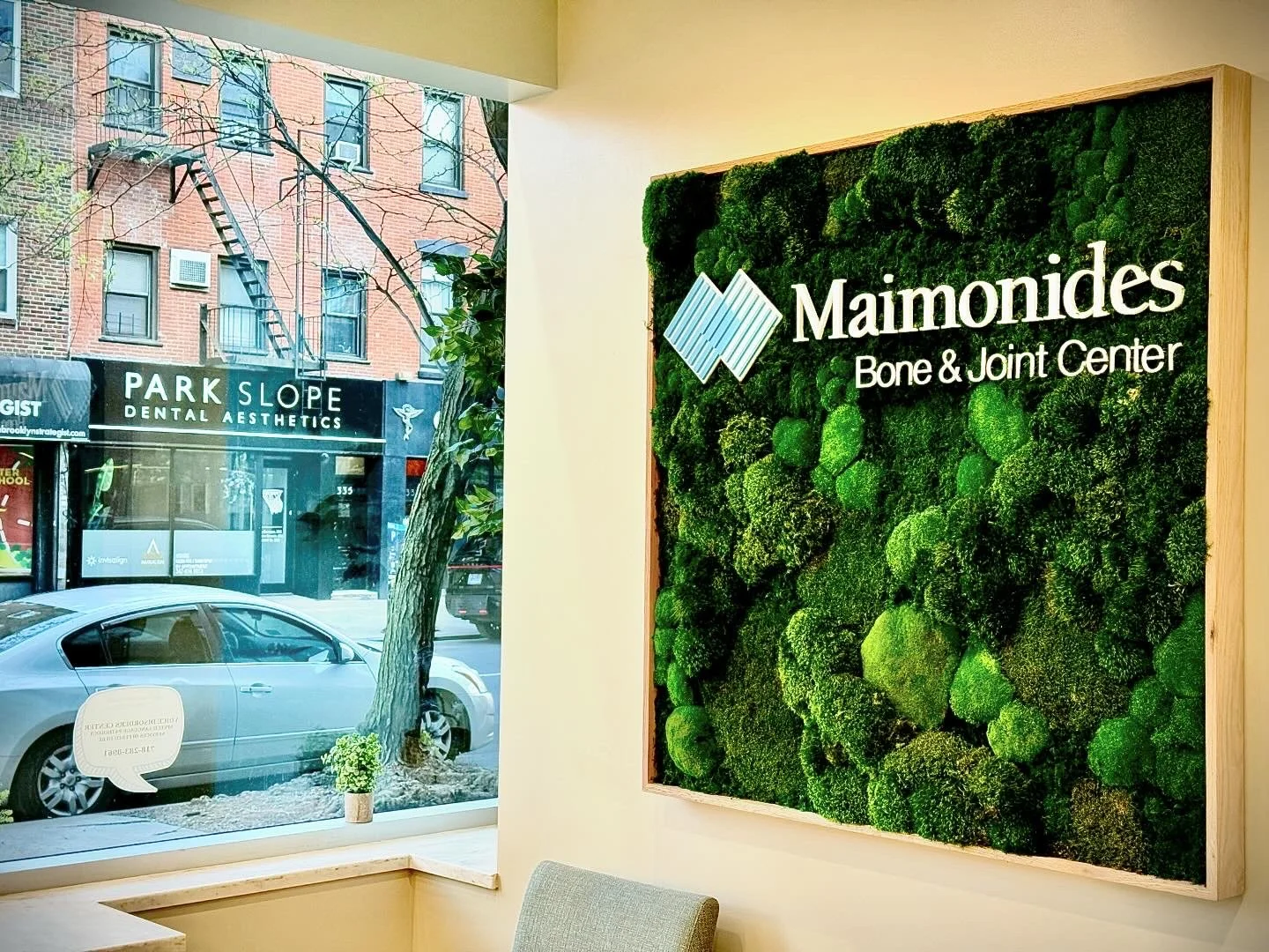 Interior of Maimonides Bone & Joint Center with a green moss wall and a window showing the street outside with parked cars and a tree.