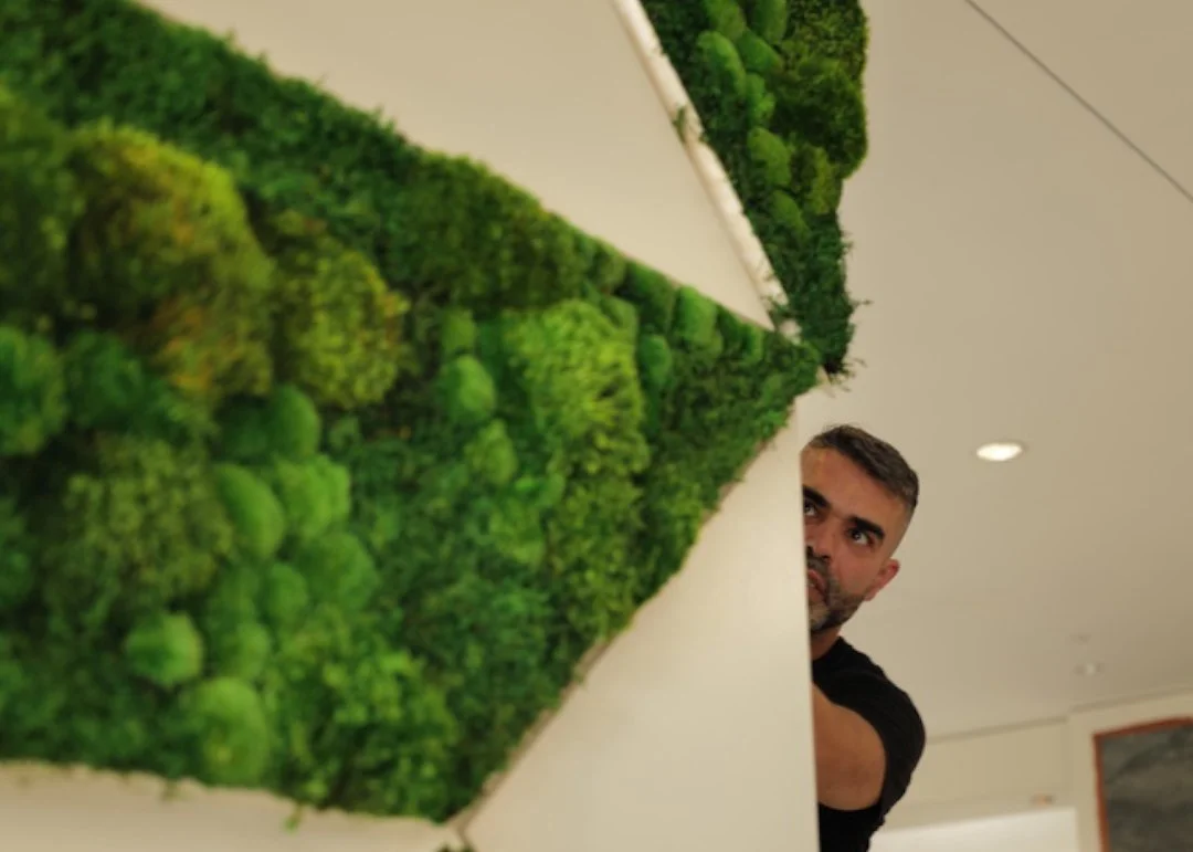 A man peeking from behind a white wall with a green moss or plant wall design.
