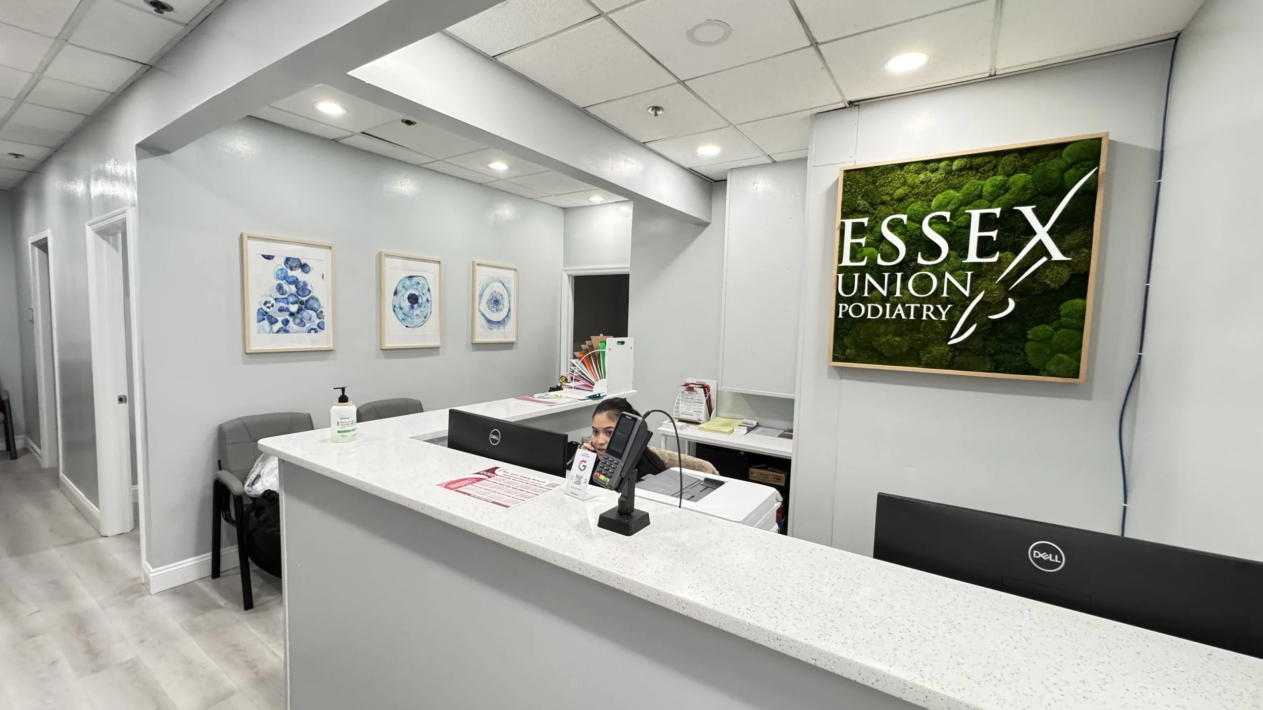 Reception area at Essex Union Podiatry with a white counter, a desktop computer, a phone, and a woman sitting behind the counter. There are framed blue artwork on the wall, a hand sanitizer bottle on the counter, and a large sign on the wall that rea