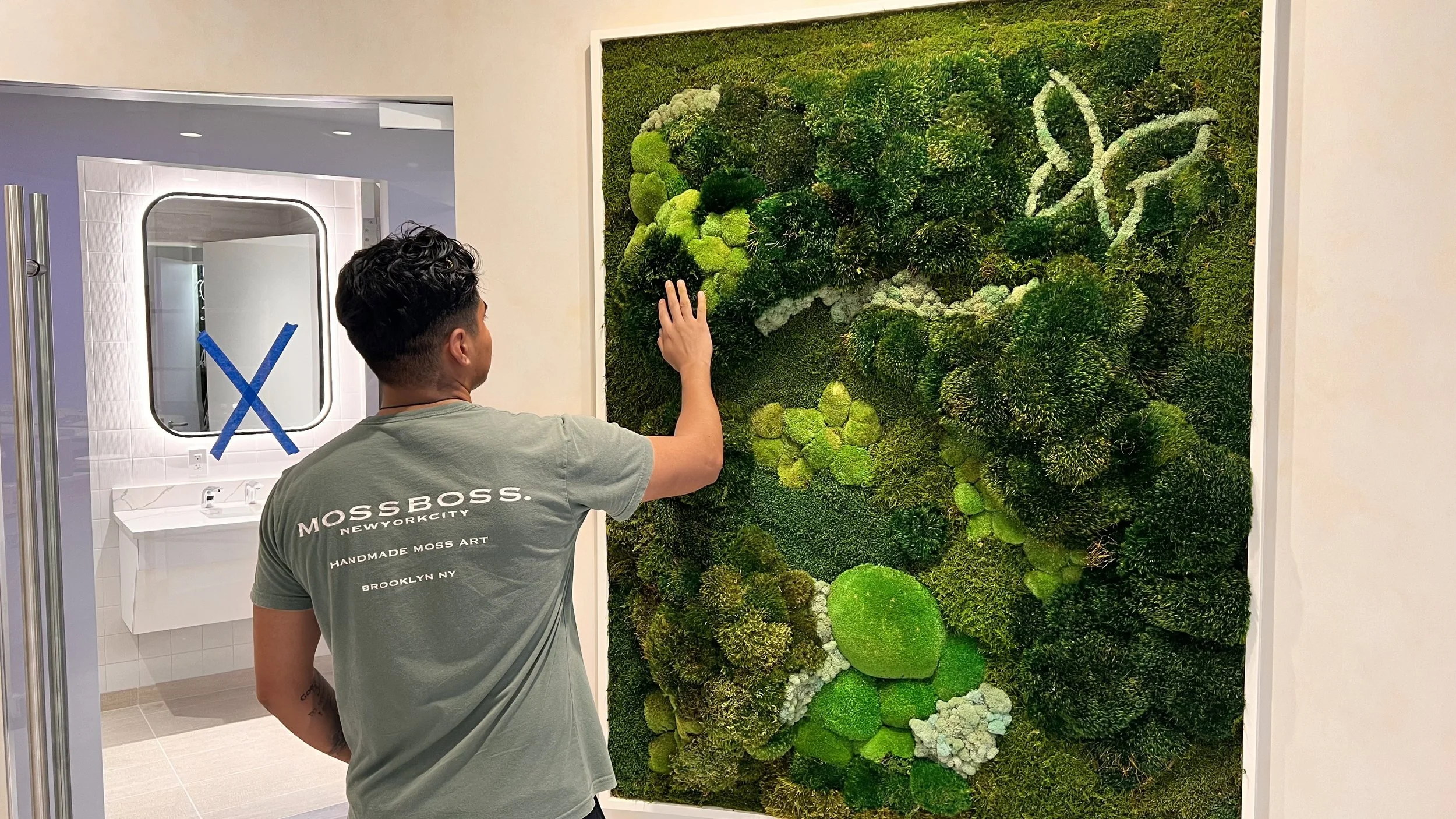A man with dark hair wearing a green t-shirt that says 'MOSS BOSS. NEW YORK CITY HANDMADE MOSS ART BROOKLYN NY' is touching a large wall art piece made of various green mosses in an indoor setting. In the background, there is a white bathroom sink wi