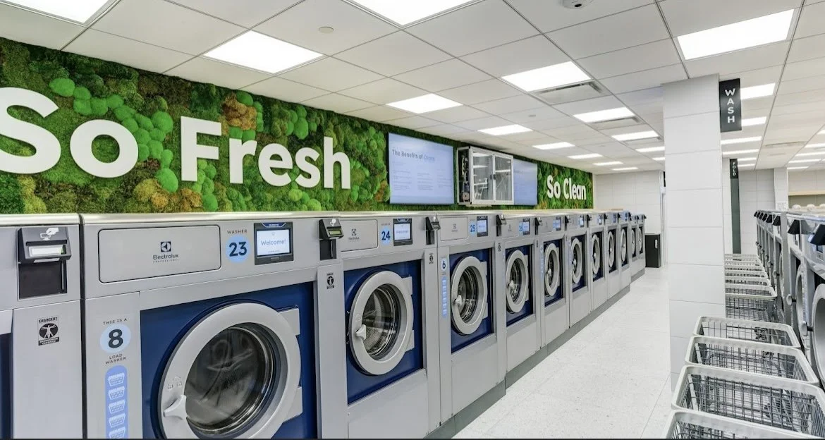 Multiple front-loading washing machines in a laundromat with signs reading 'So Fresh, So Clean' on the wall.