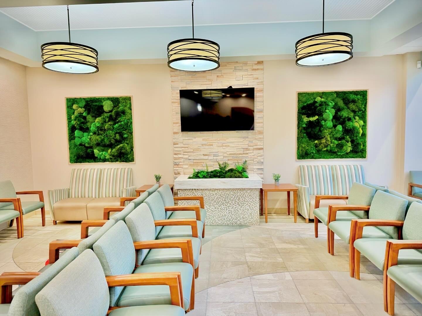 A lobby with multiple chairs arranged in a semi-circle around a corner fireplace with a plant feature above it, framed green moss artwork on beige walls, and a mounted flat-screen TV above the fireplace. Three pendant light fixtures hang from the cei