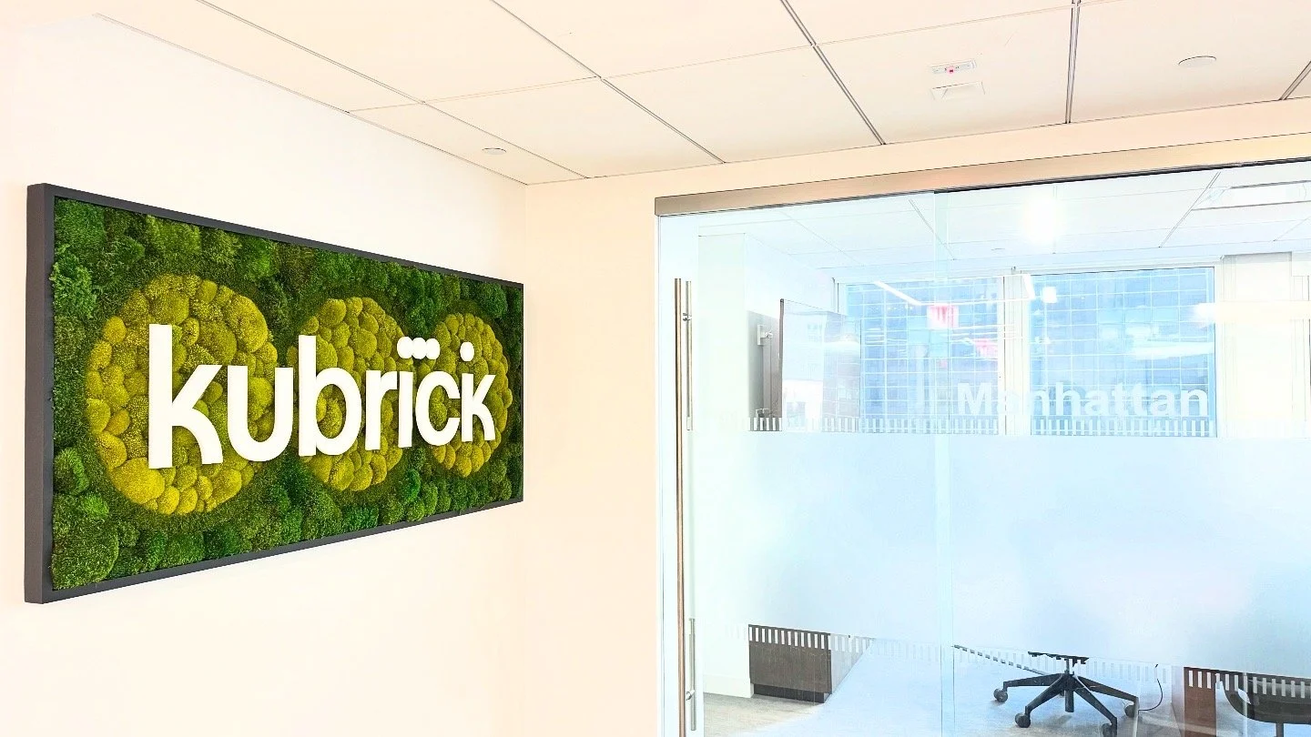Interior of an office with a large wall-mounted display showing the word "kubrick" on a green moss background, and a glass wall with city buildings and the word "Manhattan" visible outside.