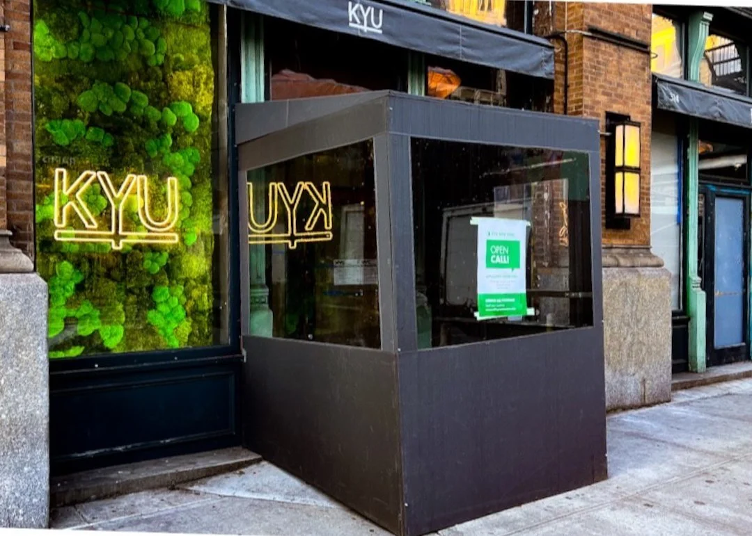 Exterior of KYU restaurant with a black entrance and neon yellow KYU signs, a green moss wall, and a black outdoor booth with a green and white open call sign displayed.