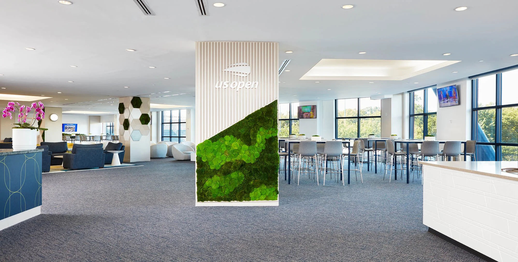Interior view of a modern lounge area at U.S. Open with seating, tall windows, and a green wall display.