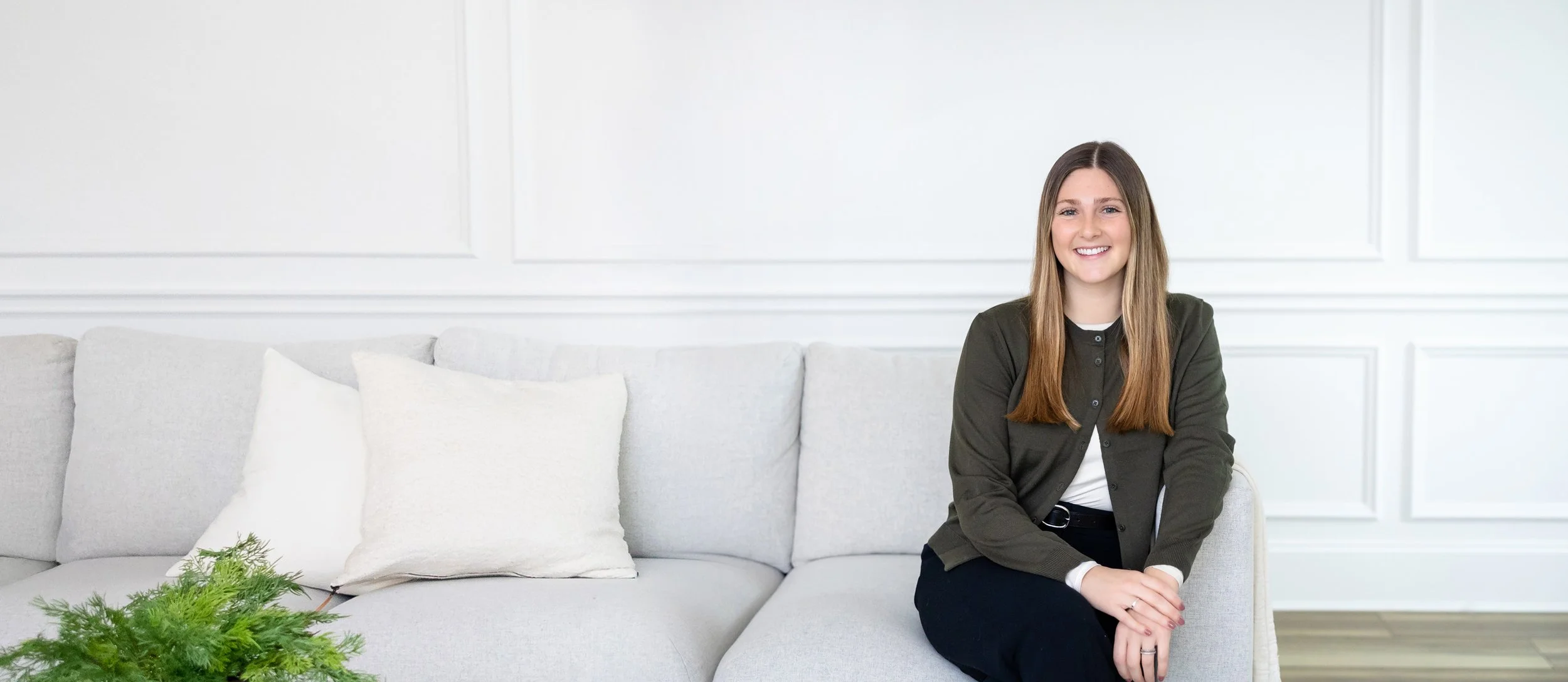 A young woman with long brown hair, wearing a dark green cardigan and black pants, sitting on a light-colored sofa with white pillows, smiling at the camera in a bright, white-paneled room.