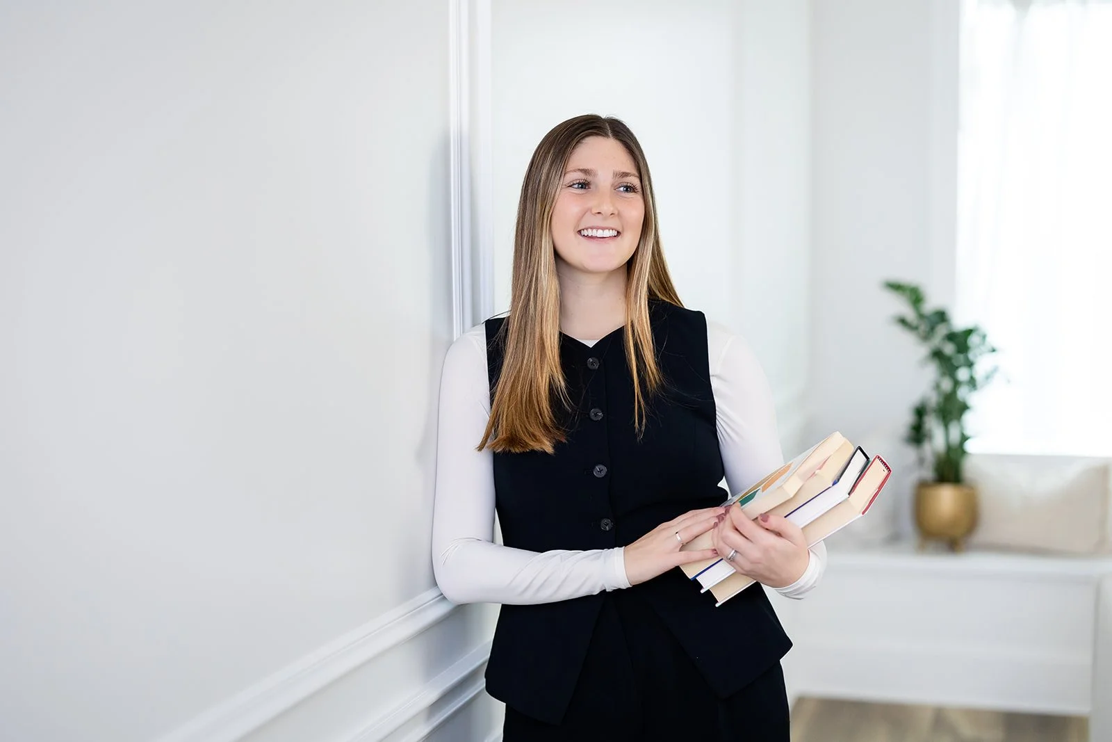 A young woman with long, straight, light brown hair, wearing a black blazer over a white shirt, standing indoors against a white wall, holding books, smiling, with a potted plant visible in the background.