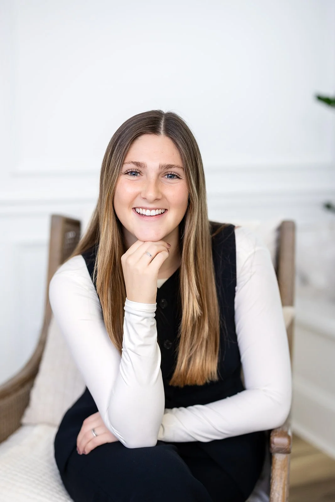 A young woman with long light brown hair smiling while sitting on a wooden chair in a bright room.