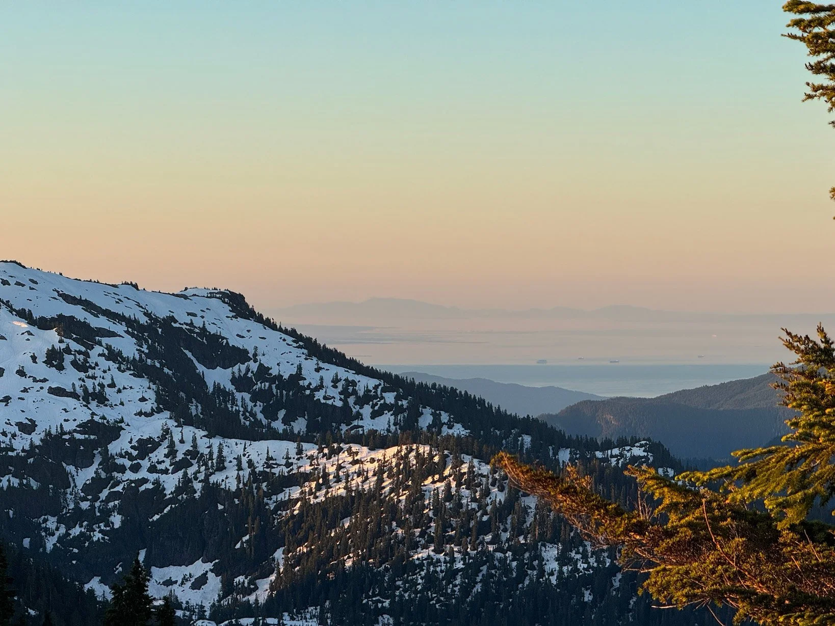 Snow-covered mountain range with evergreen trees and a distant view of water and land, under a clear sky at sunset or sunrise.
