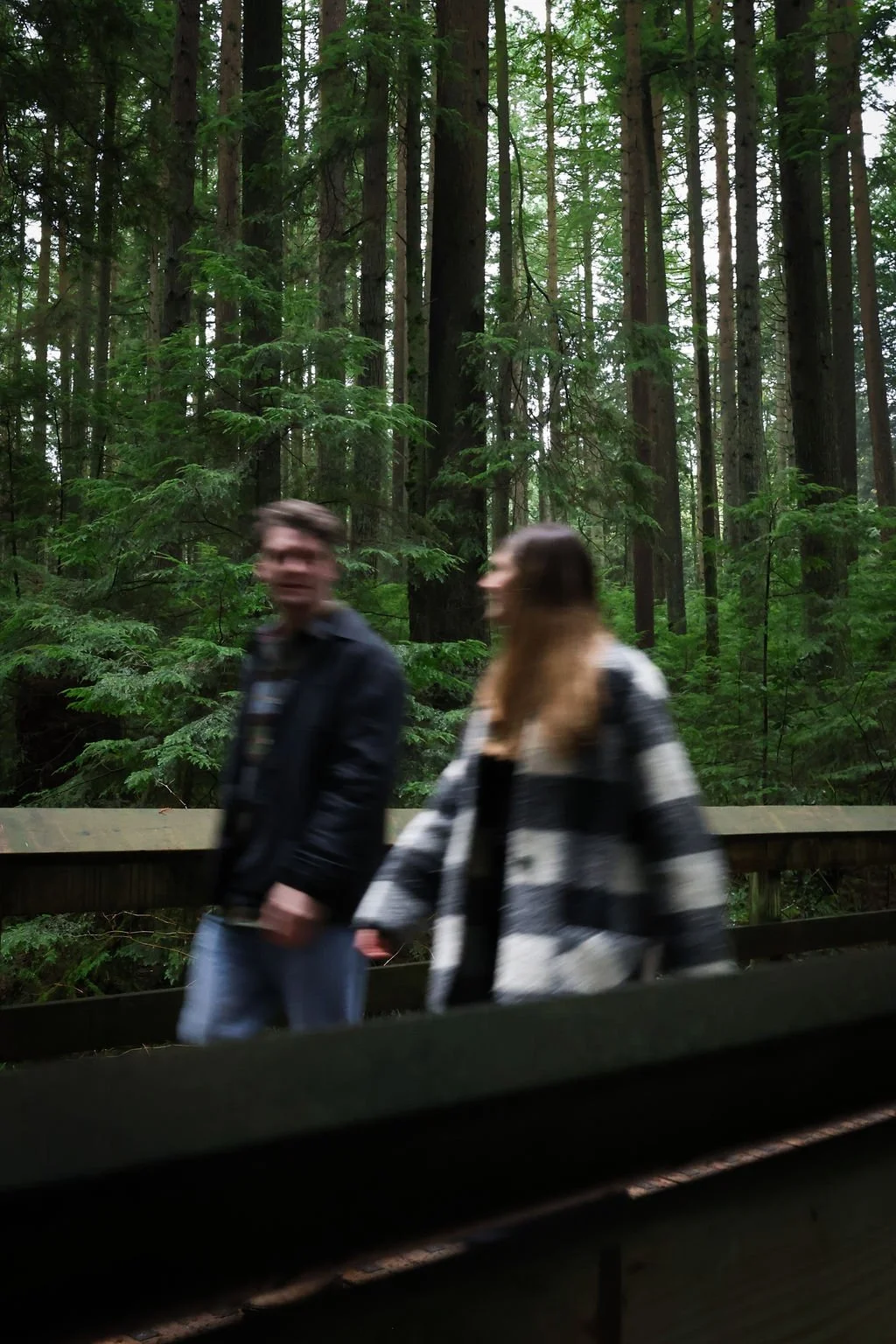 Two people walking along a wooded pathway surrounded by tall green trees in a forest.