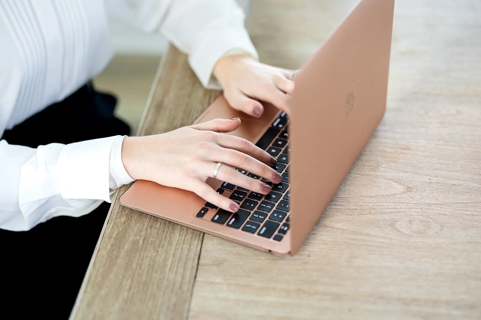 Close up of woman using a rose gold laptop on a wooden table.