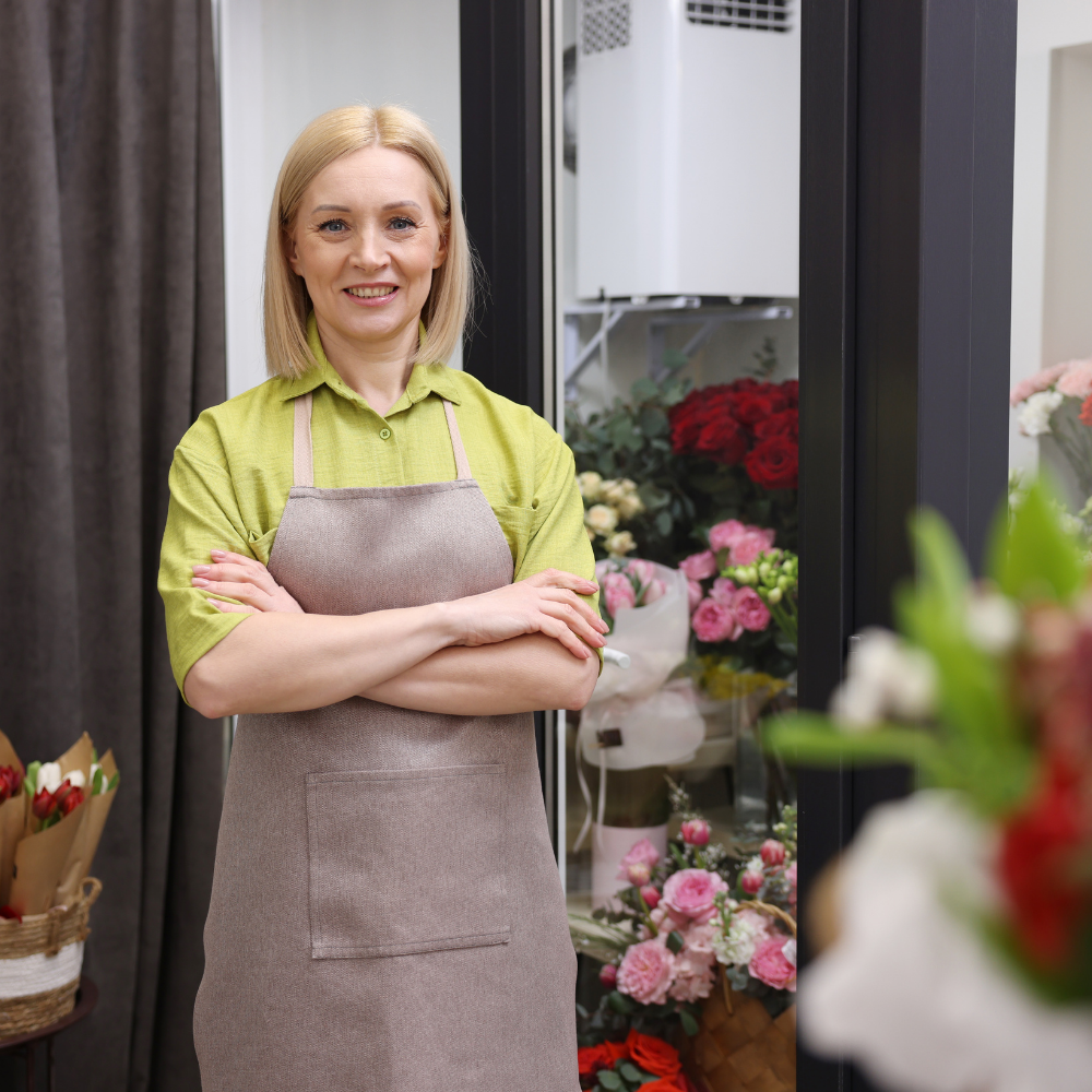A woman with blonde hair smiling and standing with arms crossed in a flower shop or floral shop, wearing a green shirt and a beige apron, with flowers in the background.