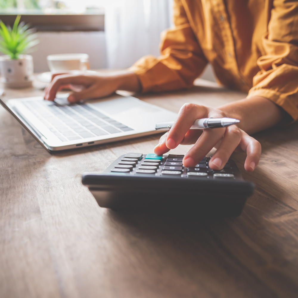 Person using a calculator with a pen in hand, working on a laptop at a wooden desk.