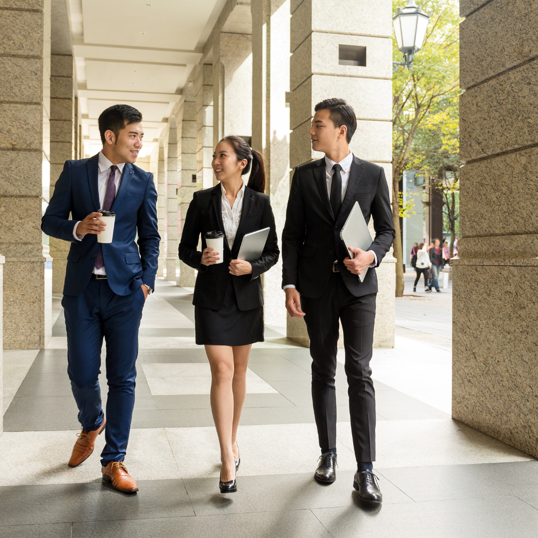 Three young professionals walking and talking outside on a city sidewalk, dressed in business attire, carrying coffee cups and folders.