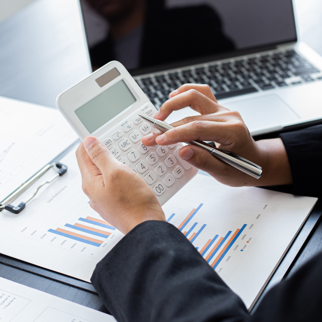 Person using a calculator with financial charts and a laptop on the desk.