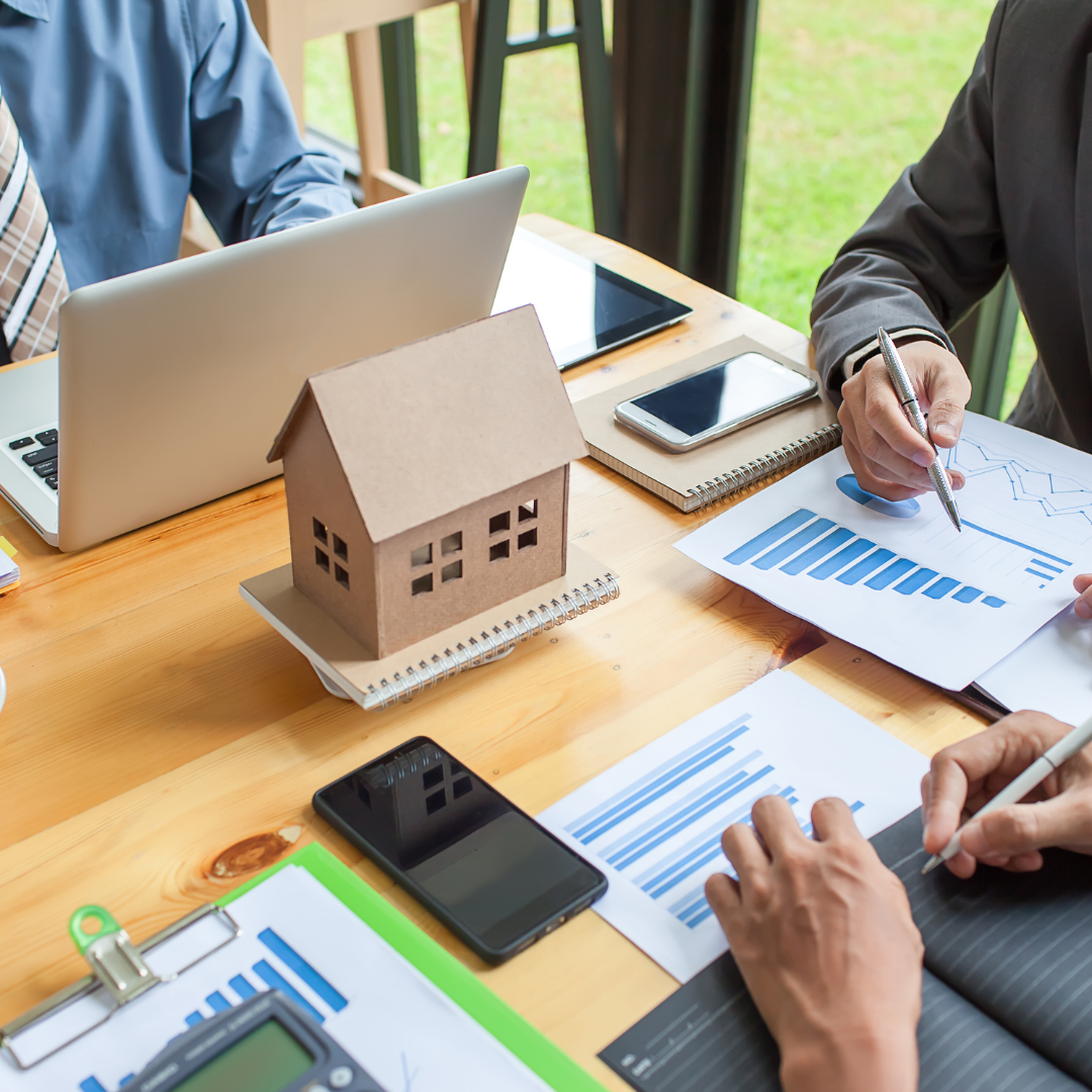 Business meeting discussing real estate investment, with papers, charts, a small house model, a laptop, and smartphones on a wooden table near windows.
