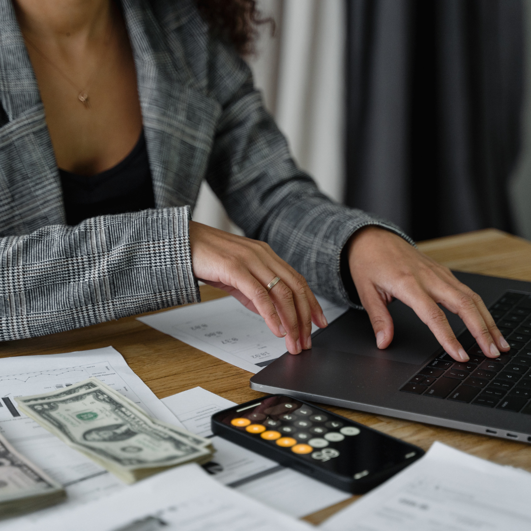 A woman wearing a checkered blazer is working on a laptop, surrounded by scattered papers, dollar bills, and a calculator on a wooden desk.