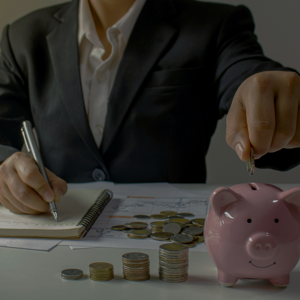 A person in a dark suit and white shirt is sitting at a table, making a financial calculation. On the table, there is a pink piggy bank with a smiley face, stacks of coins, a notepad, and some papers with financial charts.