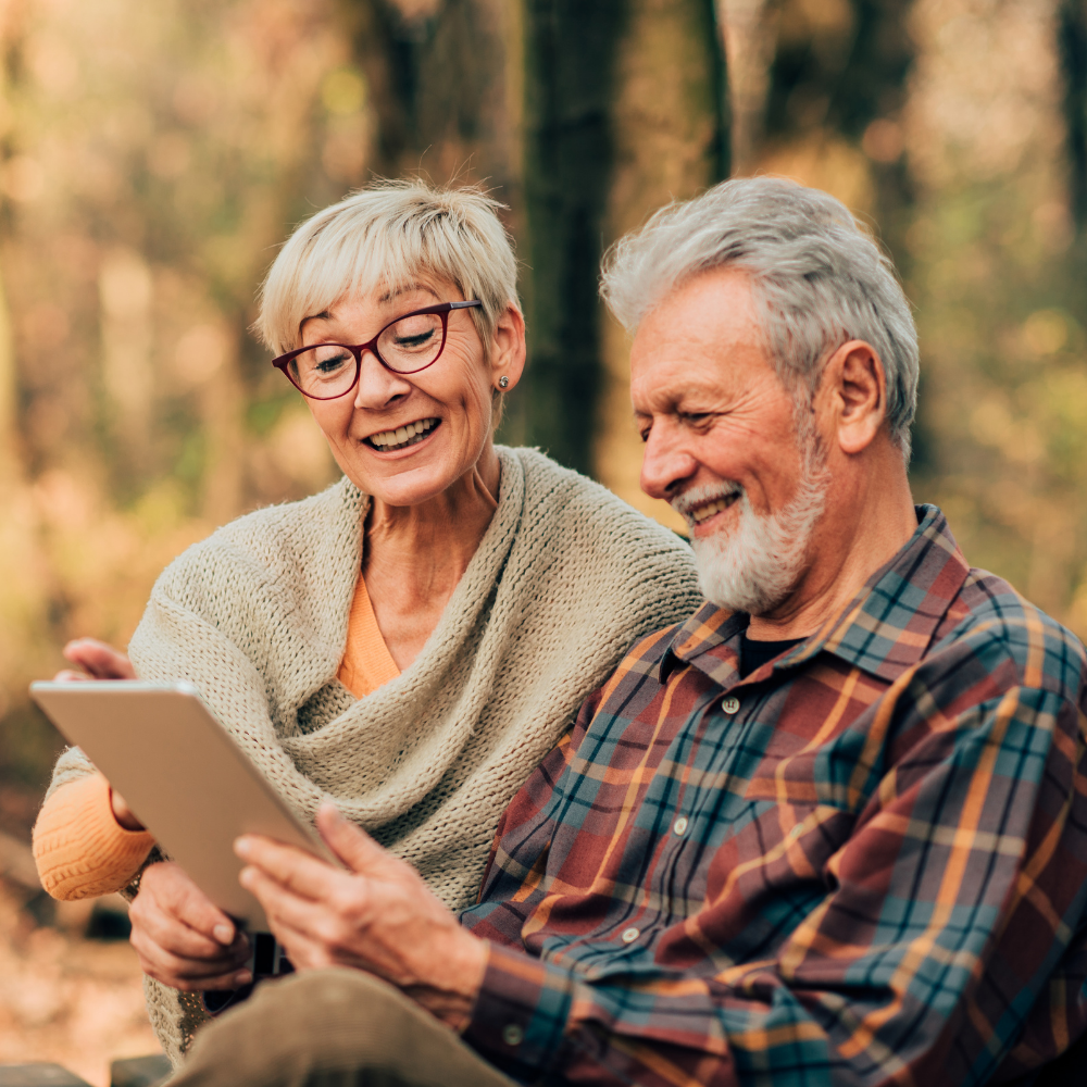 An elderly couple outdoors in a forest, smiling and looking at a tablet device.