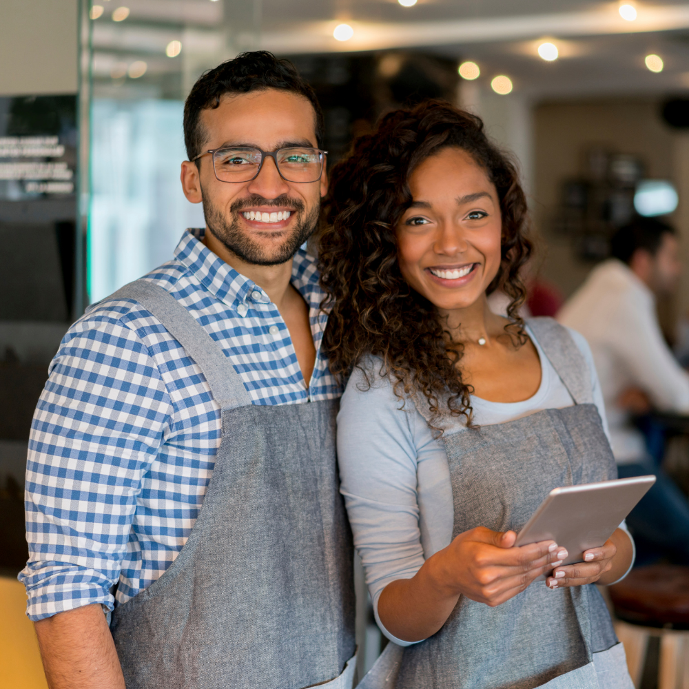 Smiling man and woman in a café, wearing aprons, with a tablet in the woman's hands, possibly waitstaff.