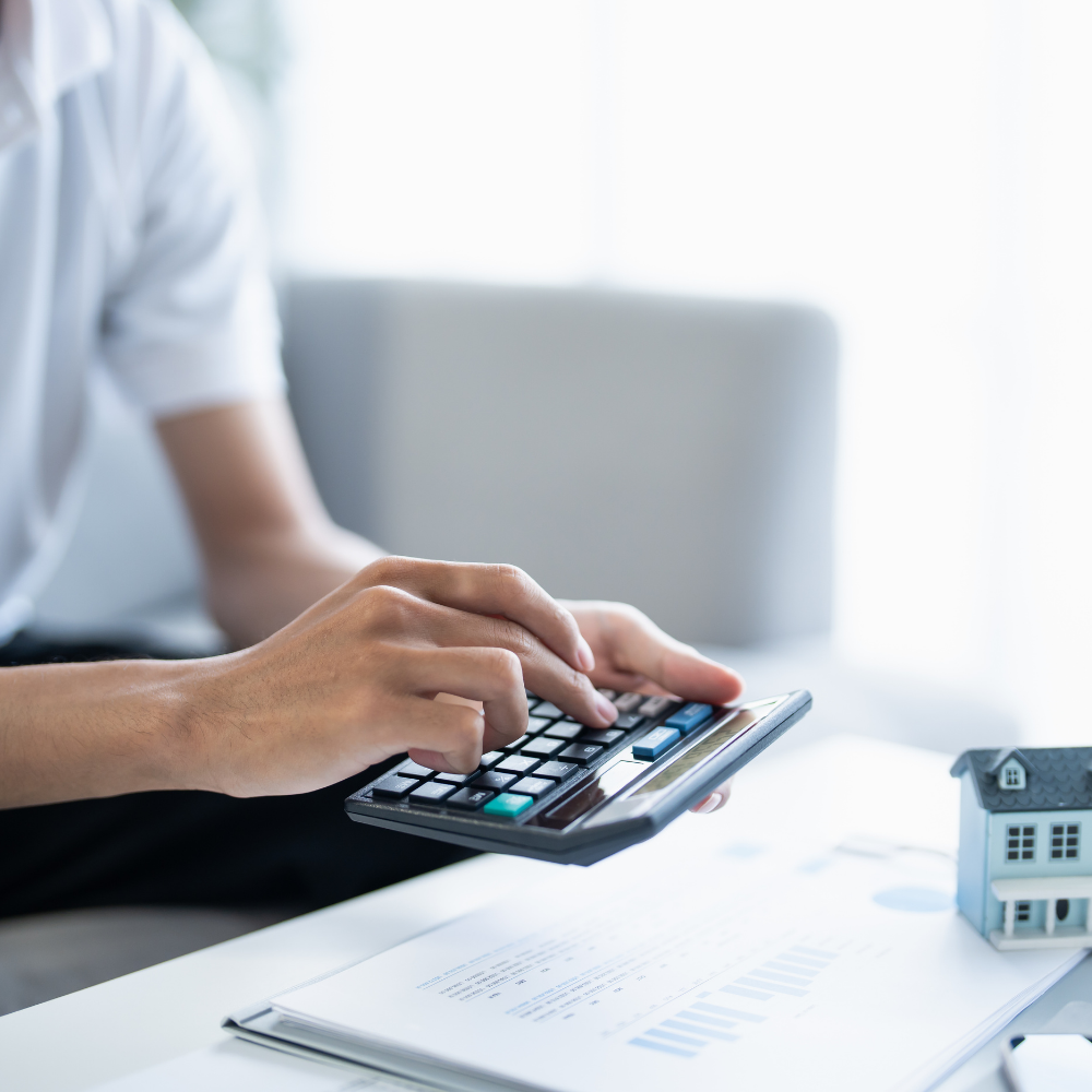 Person using calculator on desk with papers and small house model.