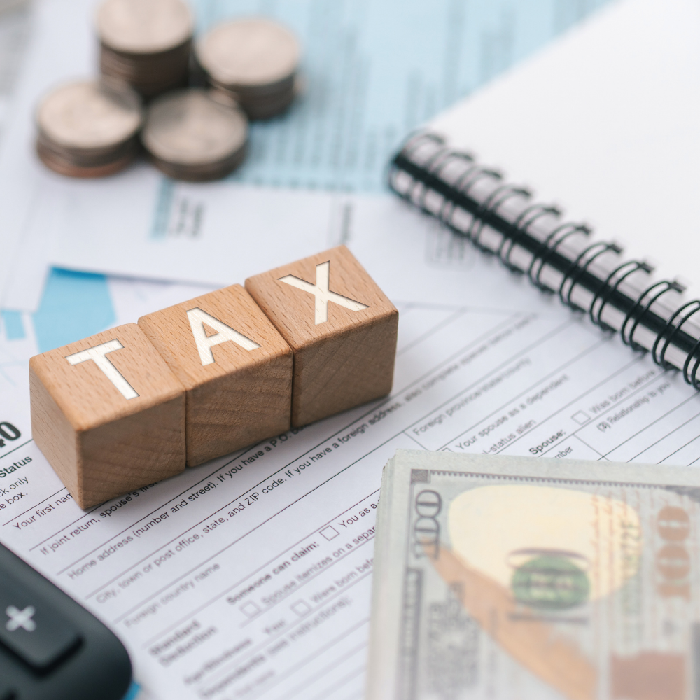 Wooden blocks spelling 'TAX' placed on a desk with financial documents, a spiral notebook, a calculator, U.S. hundred-dollar bills, and coins.