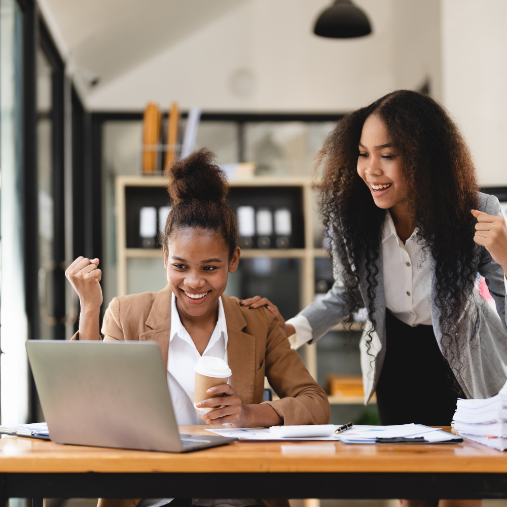 Two women celebrating success at a work desk with a laptop, papers, and coffee in an office setting.