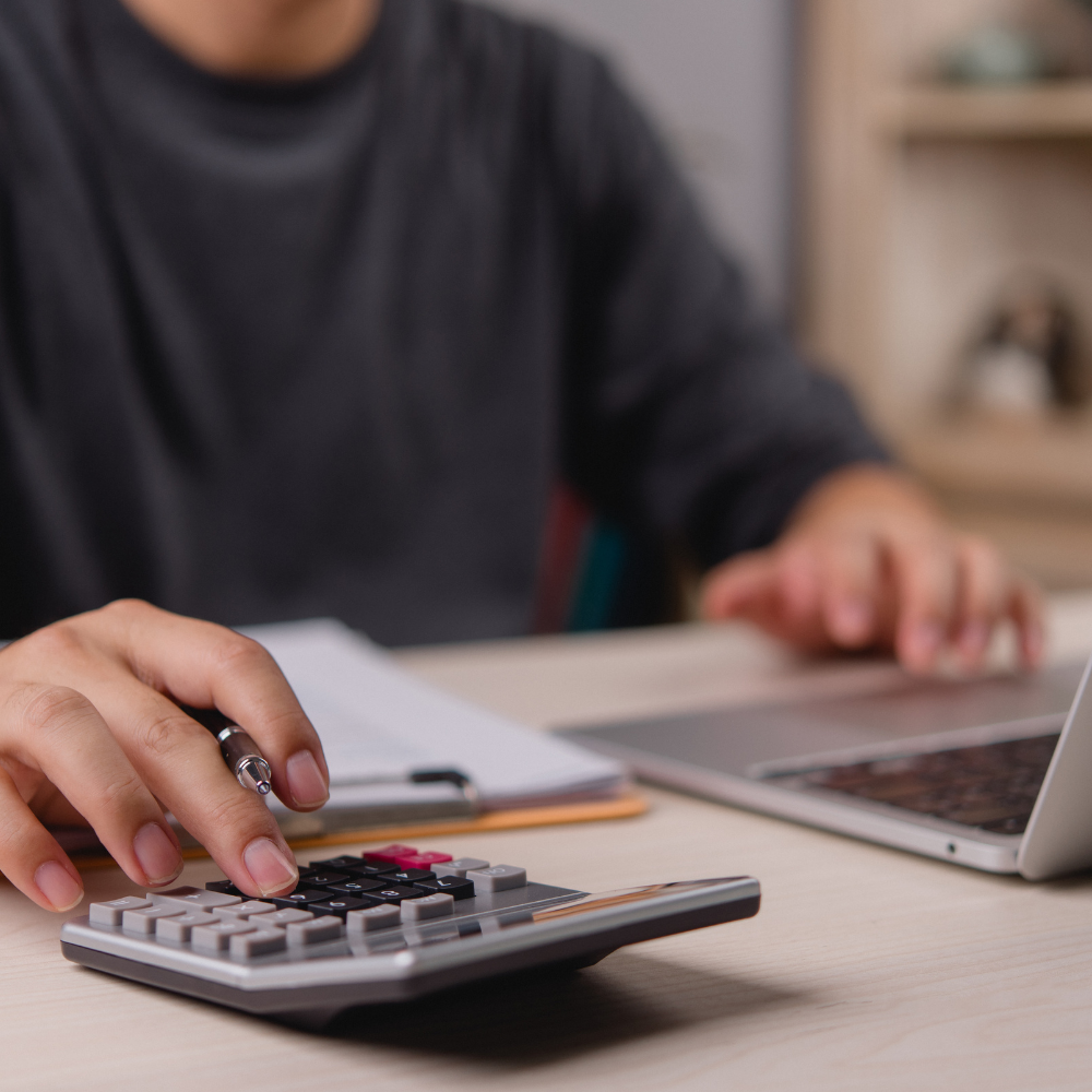 Person using a calculator with a pen, laptop, and notebook on a desk.