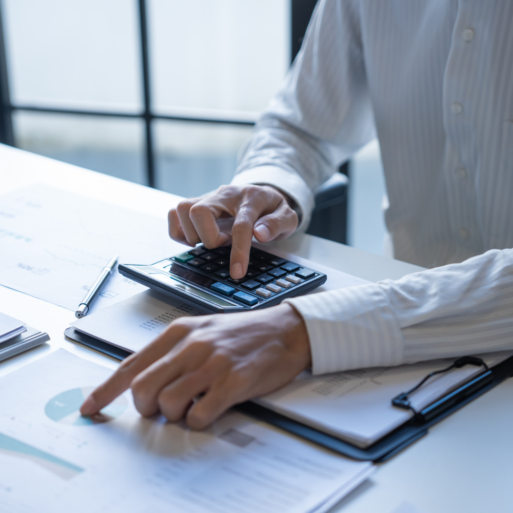 Person wearing a white striped shirt working with a calculator, documents, and a pen on a desk with a window in the background.