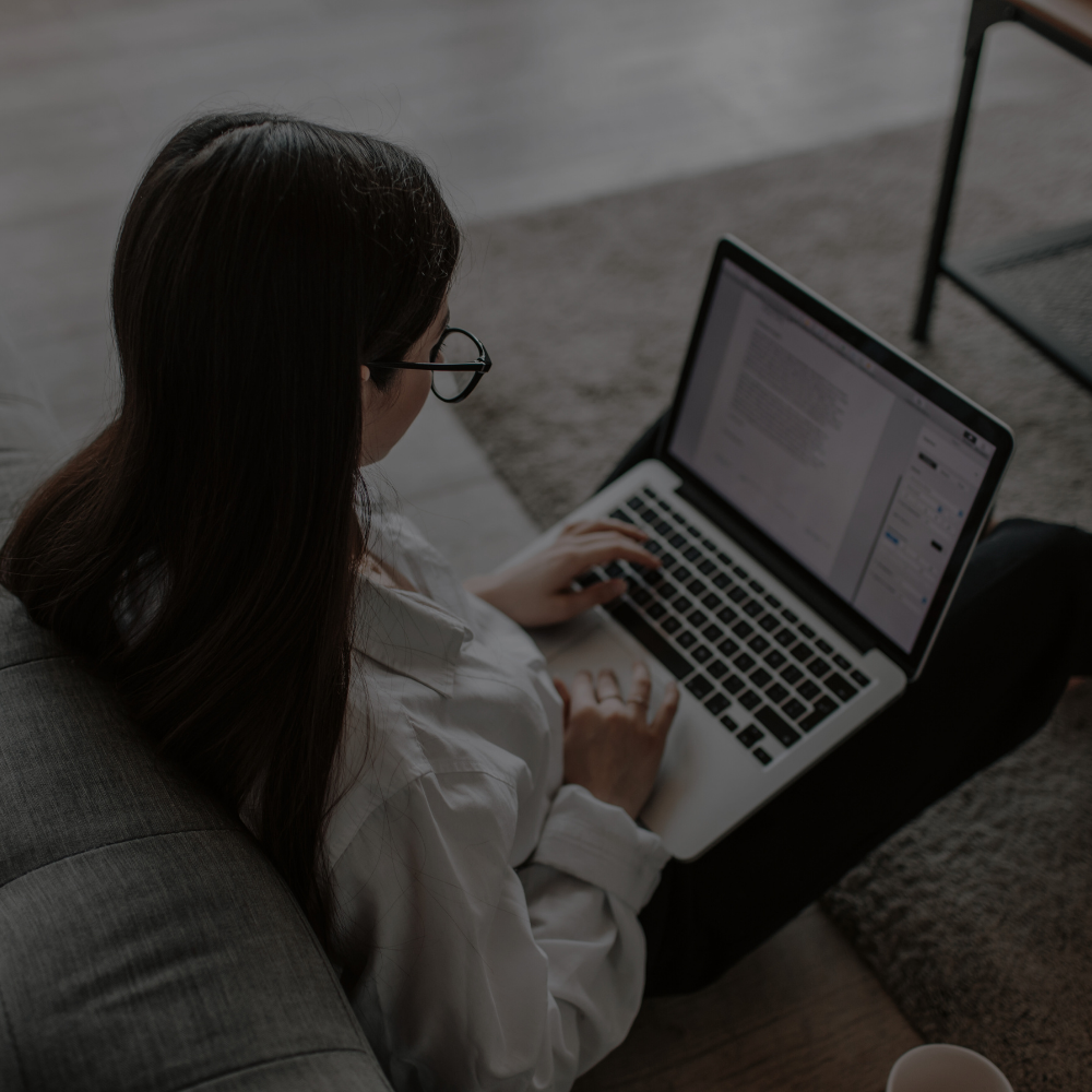 A woman with long dark hair and glasses sitting on a gray sofa, working on a laptop with a document open on the screen, in a cozy indoor setting.