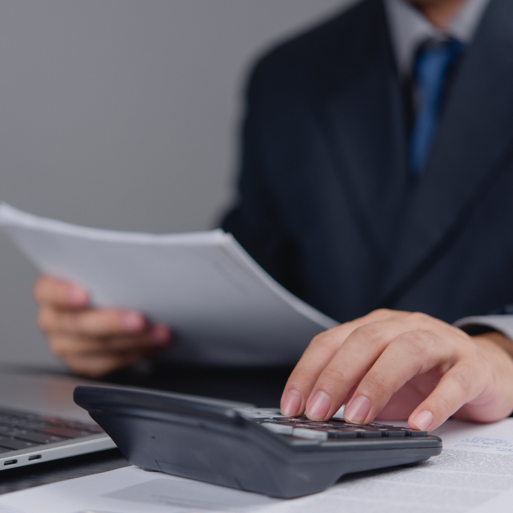 A person in a suit using a calculator with papers and a laptop on a desk