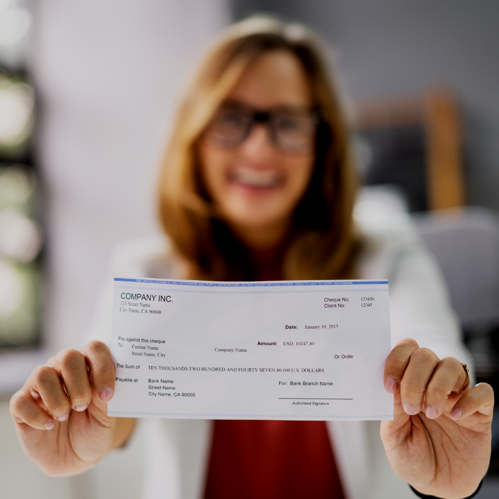 A woman with glasses and long hair smiling and holding a check in front of her.
