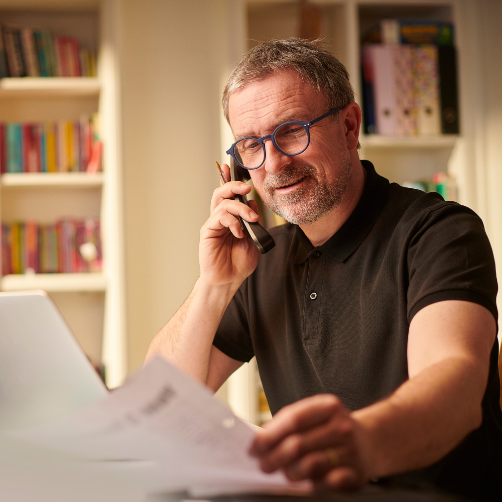 Man with glasses talking on the phone while reviewing papers at a desk in a room with bookshelves.