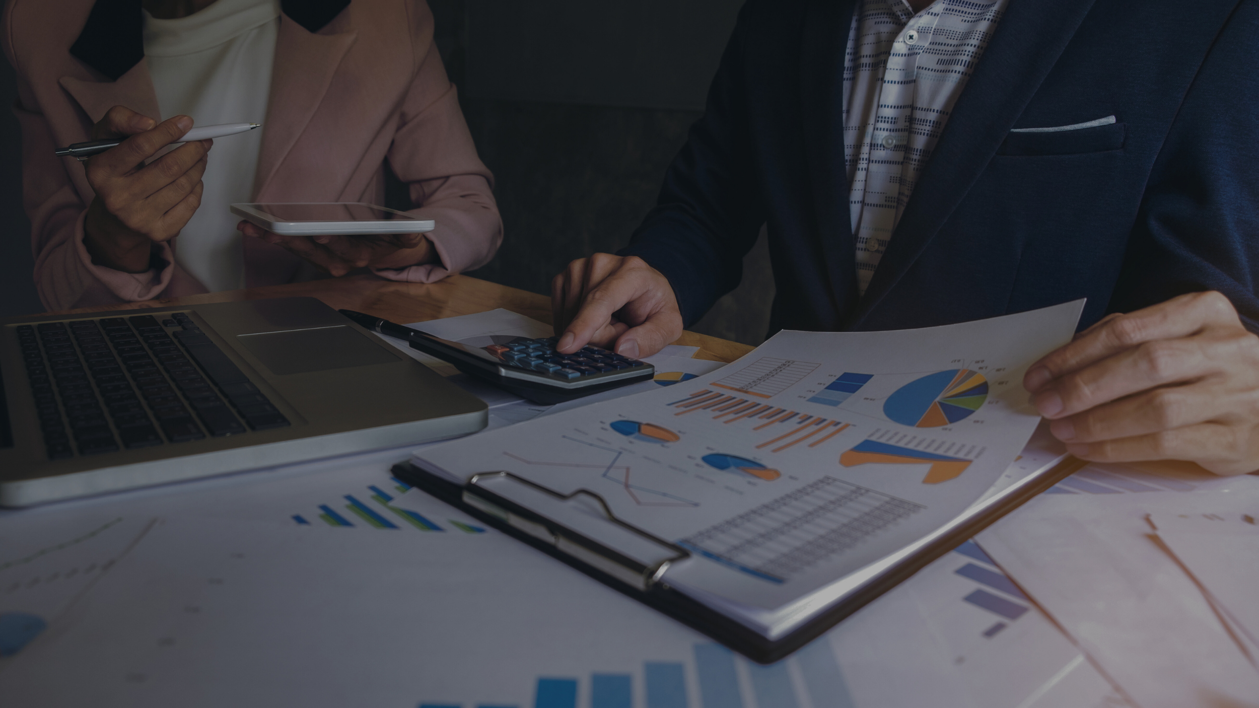 Two people are sitting at a desk with financial documents, using a laptop, calculator, tablet, and pen, analyzing charts and graphs.