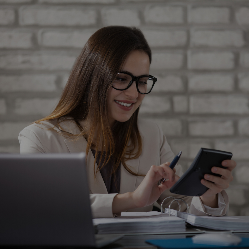A woman with brown hair, wearing glasses and a beige blazer, is sitting at a desk with a laptop, papers, and a calculator, smiling while looking at her phone.