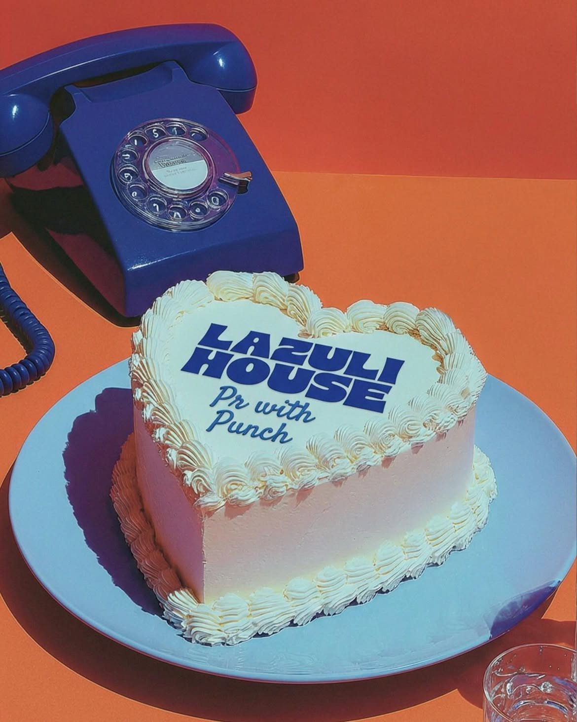 A vanilla heart shaped cake has the Lazuli House logo plus "pr with punch" written underneath in icing. A blue retro phone in the background sits on a papaya-colored table.