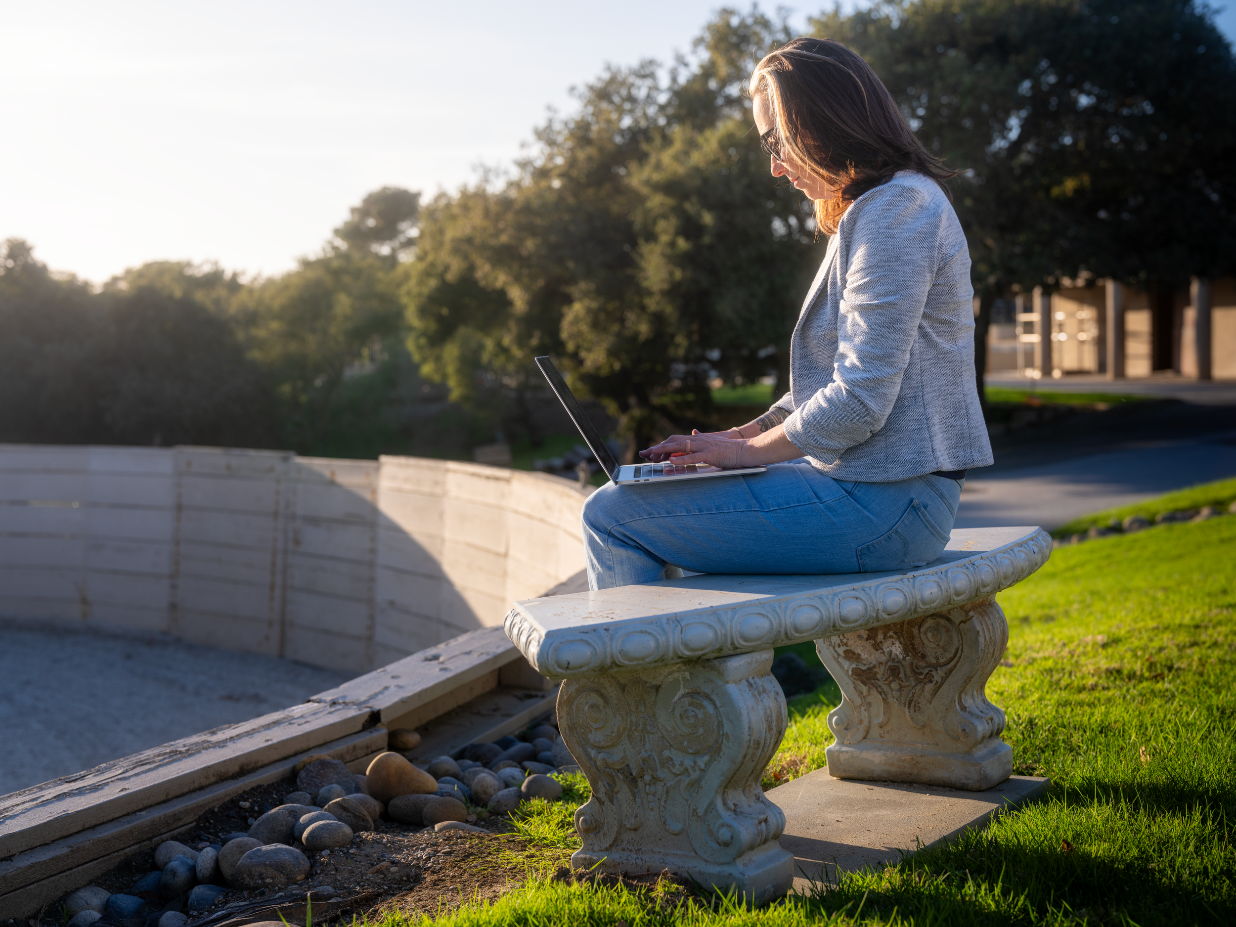 A woman sitting on a stone bench outdoors using a laptop, with trees and a building in the background during sunset.