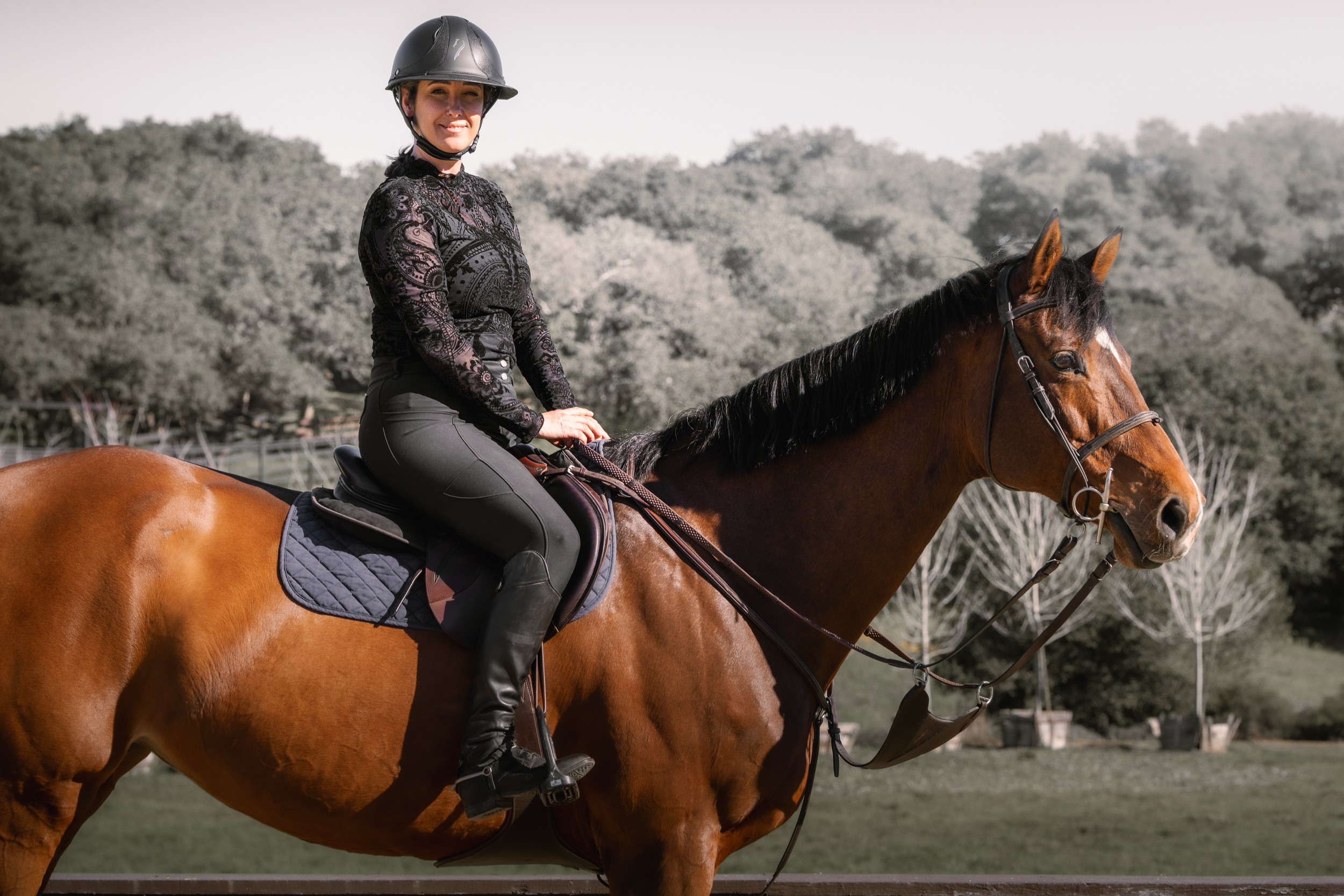 A woman riding a brown horse outdoors with trees in the background.