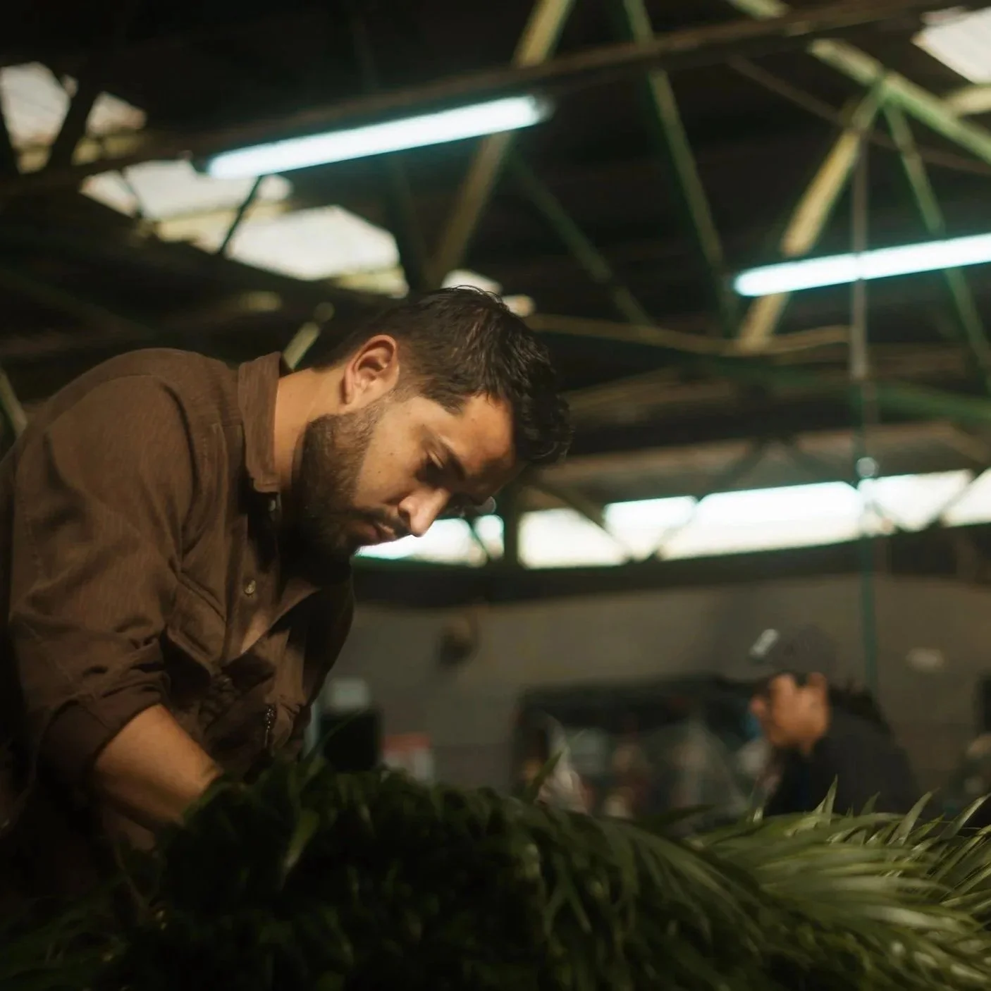 A man with a beard, wearing a brown jacket, looking downward and focused on something in front of him inside a workshop or studio with a high ceiling and exposed metal beams; another person is in the background.