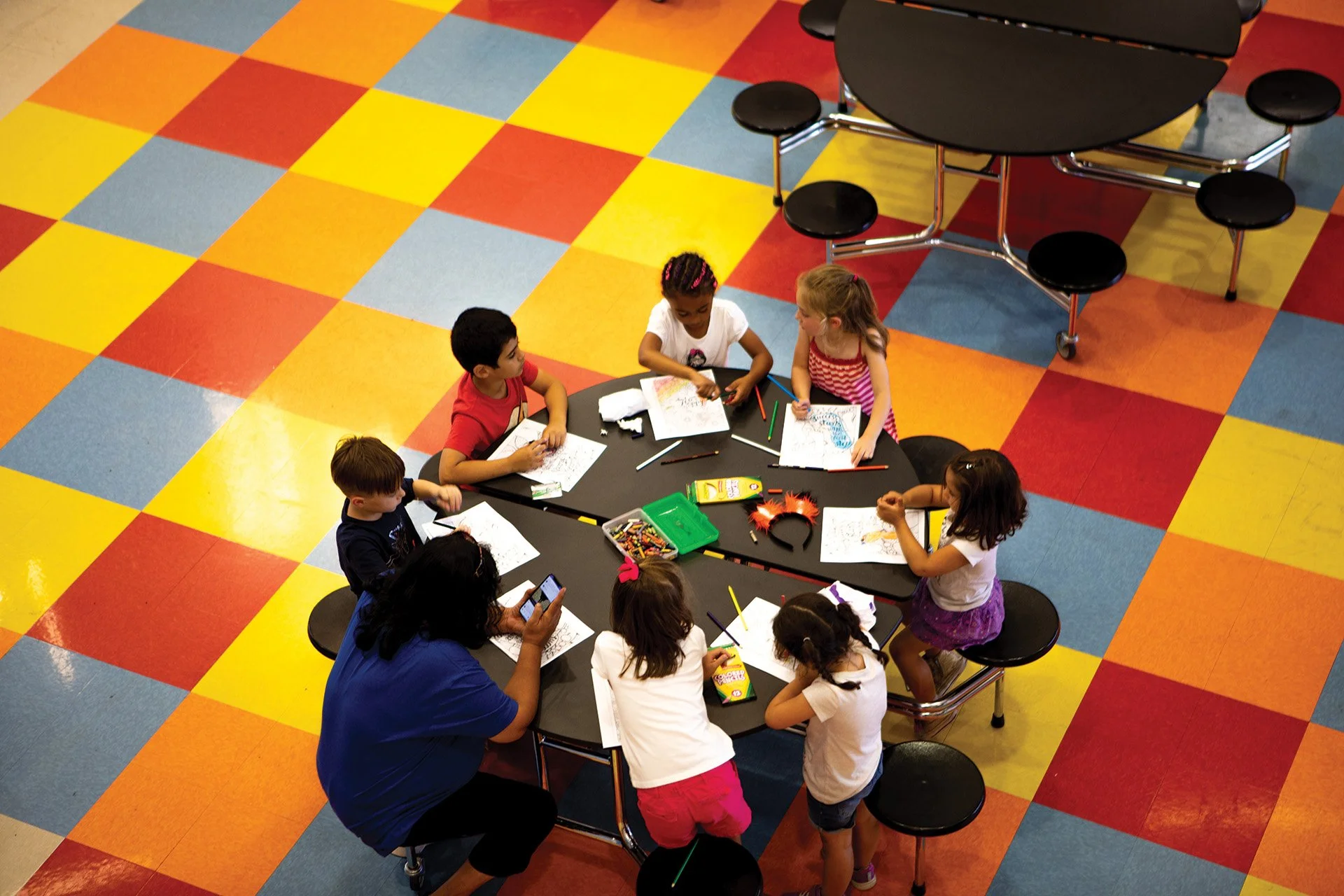 Children and an adult sitting around a table in a colorful room, engaged in coloring activities. The room has bright yellow, red, blue, and orange checkered flooring.