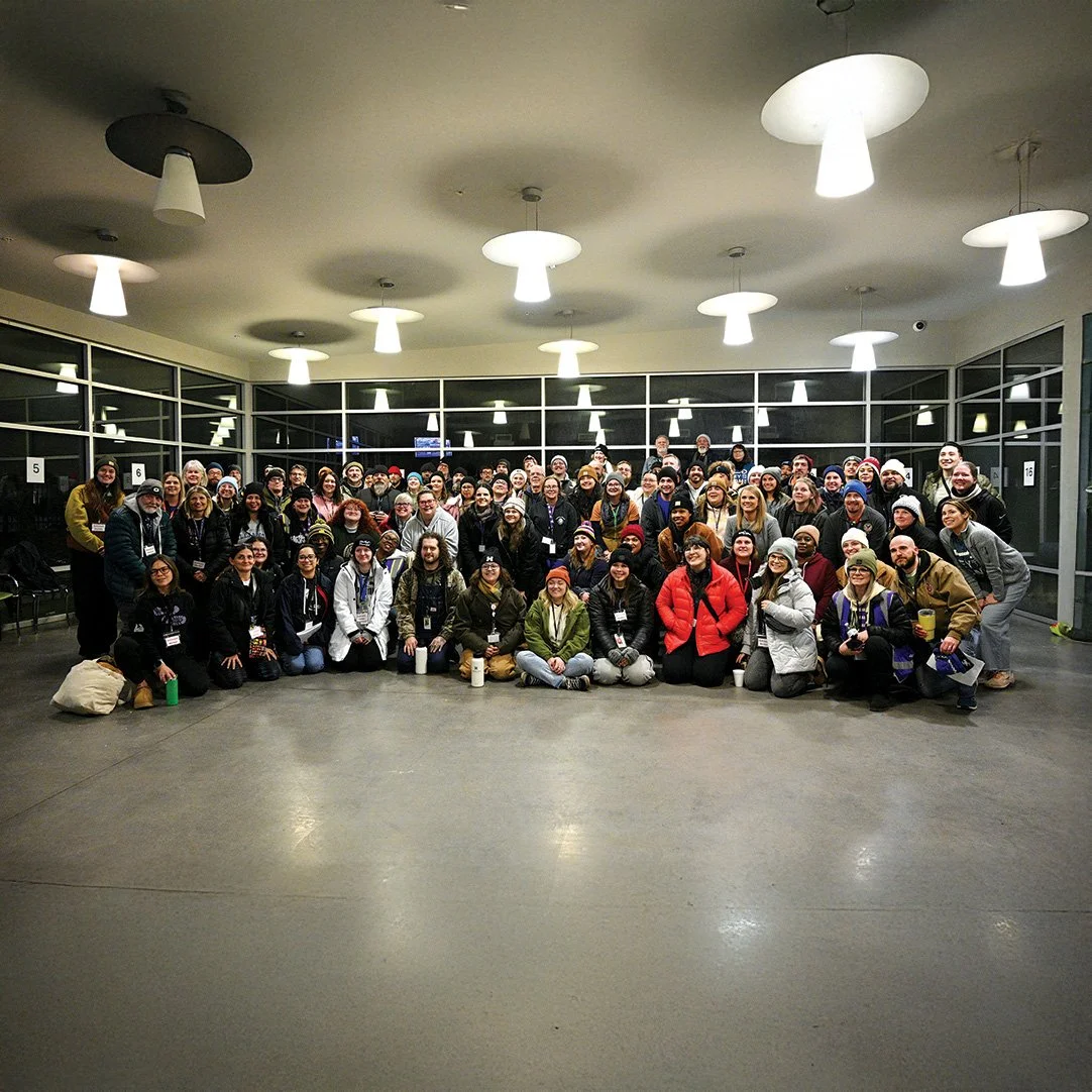 Large group of people gathered indoors in a well-lit room with large windows, posing for a group photo.