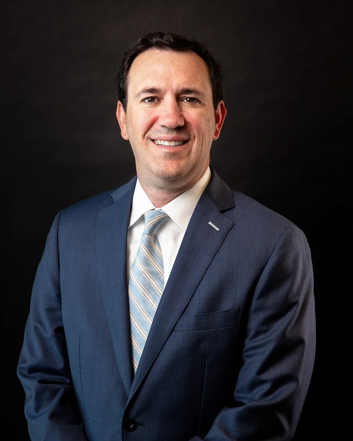 A professional headshot of a man in a suit smiling against a gray background.