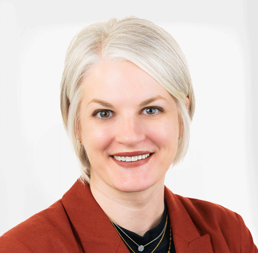 A woman with short blonde hair, blue eyes, and light skin, wearing a rust-colored blazer and jewelry, smiling against a white background.
