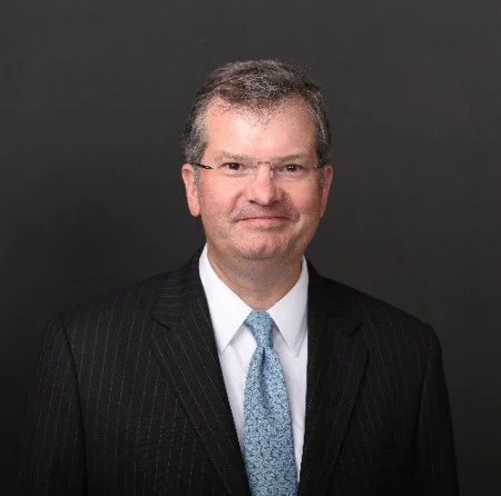Headshot of a middle-aged man in a dark suit and tie against a black background.