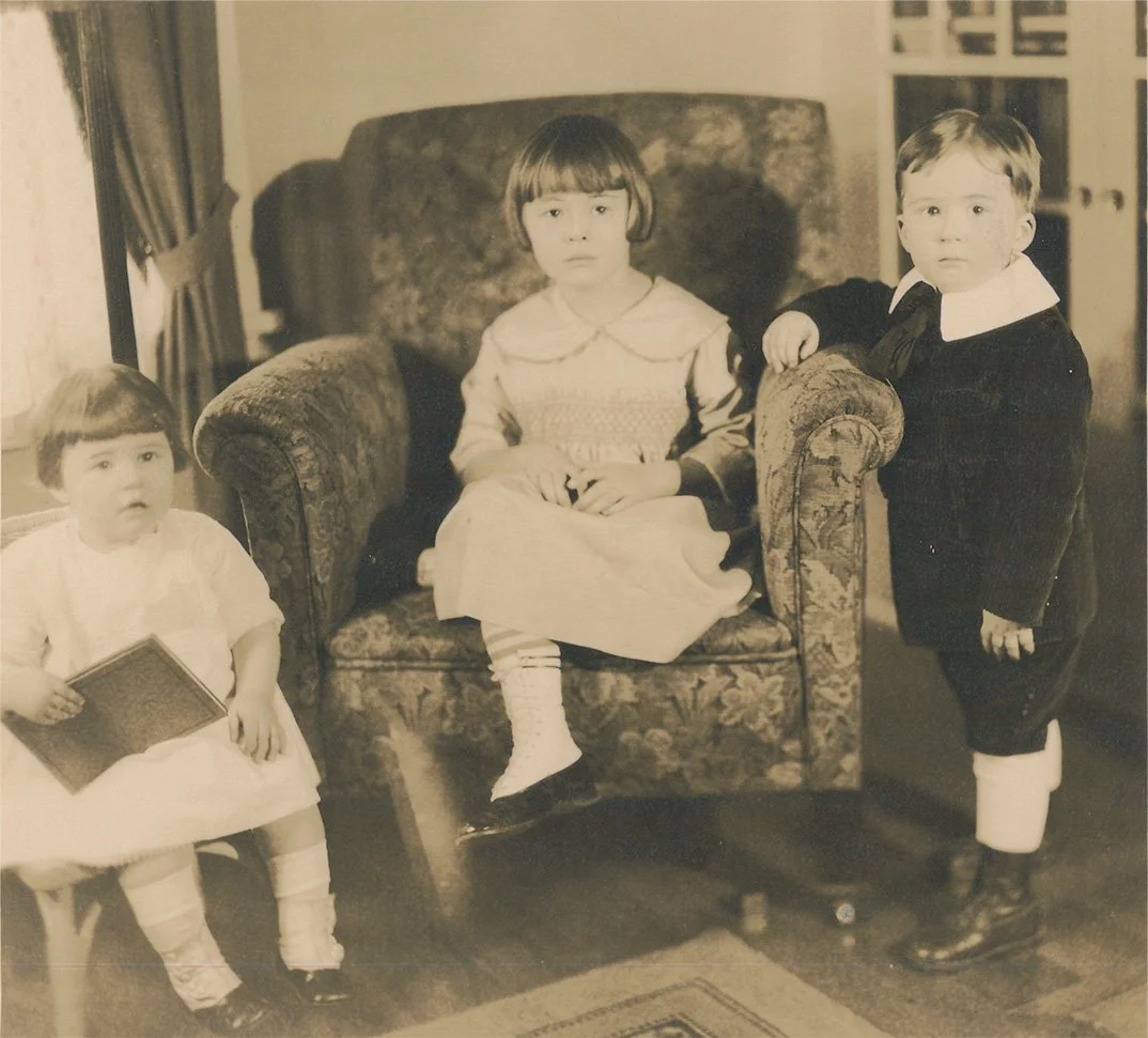 A vintage black and white photo of three children indoors. One girl is sitting on a floral armchair, another girl is sitting on a chair holding a book, and a boy is standing beside the armchair with his hand resting on it, all dressed in old-fashioned clothing.